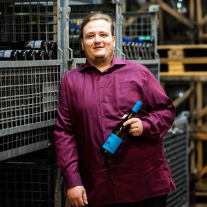 Alexander Heinrich steht vor einer Gitterbox und hält eine Flasche Wein